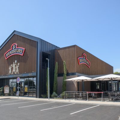 Ce restaurant 3 Brasseurs, situé à Vitrolles, est célèbre pour ses bières artisanales dans une ville dynamique des Bouches-du-Rhône. Le style du bâtiment, en forme de grange, est constitué d'une structure mixte bois / métal. Les bardages sont mis en valeur grâce aux tôles pliées imitant des profils métalliques.